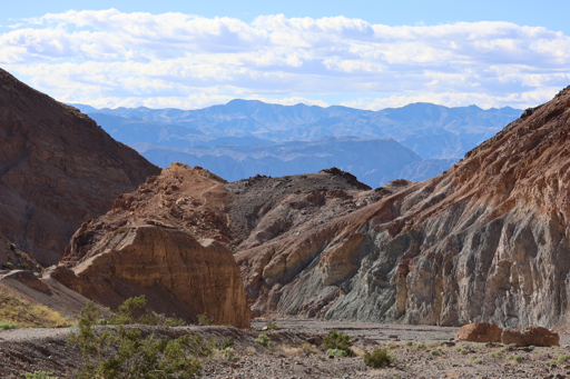 Looking back out of the Mosaic Canyon, multi-colored cliff walls may be seen on both sides with a wide gravel filled wash in the middle. Distant mountain ranges may be seen fading nto blue coloration under a bed of white clouds.