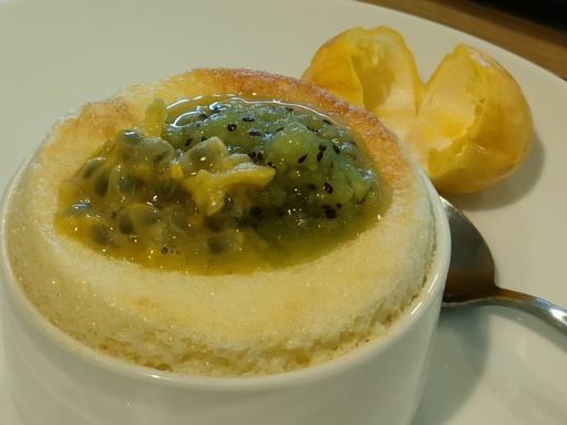 Delicious looking passionfruit souffle topped with yellow passionfruit and kiwifruit pulp in a white souffle dish on a white side plate with a teasoon to the side and a split open yellow passionfruit shell