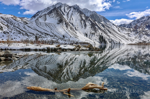Looking across the mirrored finish of Convict Lake, with Mono Jim Peak (and Mt Morrison directly behind it). The still waters perfect reflectly the image as well as allow the gravel bottom to be seen through them. A small islet covered in snow sticks out into the middle of frame and there is also a log laying half submerged in the bottom of frame.