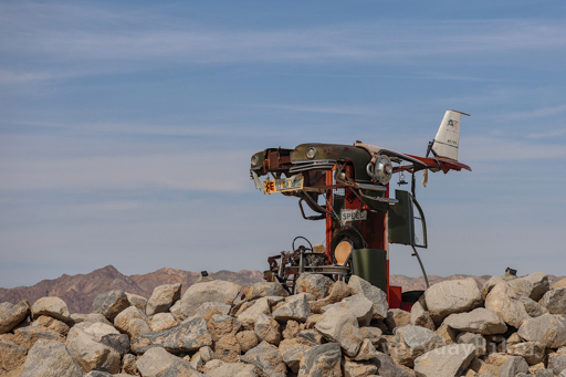 An interesting sculpture of a 50s style car converted into a small floating plane. It is surrounded by small boulders and in the distance a mountain range may be seen. License plates cut and pieced together form the word 'REINCARNATION.'