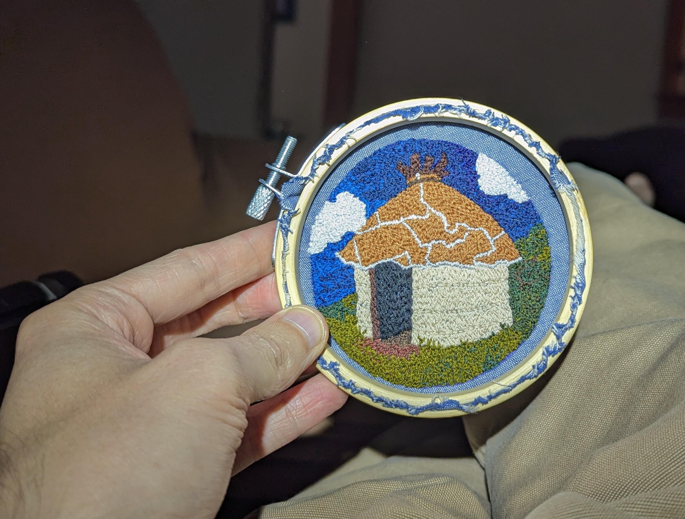 A hand holding up an embroidered patch, which is still in the embroidery hoop. It's of an Australian aboriginal style hut, with green grass and blue skies around.